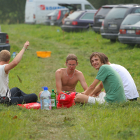 Four young men sit on the grass in a parking area between rows of parked cars, reminiscent of a 2011 summer gathering. They are gathered in a circle with some belongings, including plastic bottles and bags, in the middle. One of them, shirtless, smiles directly at the camera while another, wearing a green shirt, also smiles.