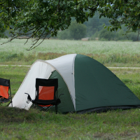A green and white tent is set up in a grassy area between two trees, with a field in the background. Two camping chairs with orange backs are positioned in front of the tent, facing each other. The serene outdoor camping setup on this cloudy day evokes memories of Laba Daba 2011.
