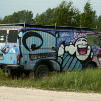 A vintage van, painted with colorful graffiti, sits on the grass. The artwork features stylized faces, abstract shapes, and vibrant colors like blue, purple, and white. Trees and a gravel road are visible in the background. The van's license plate reads "FD-7361 Laba Daba," marking its unique 2011 addition.