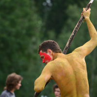 A person with their back to the camera is covered in yellow body paint with red paint on their face. They are holding a large stick above their head, poised to strike downward. The backdrop of blurred figures and green foliage hints at an outdoor setting, possibly from a 2011 Laba Daba festival.
