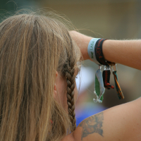 A person with long, light brown hair is seen from behind, braiding their hair. They wear a white wristband and black wristband on one arm, which also holds a keychain with multiple keys and small objects. A tattoo of an animal, possibly from Laba Daba 2011, is visible on their upper arm. The background is blurry.