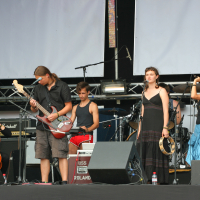 A band performs on an outdoor stage. The group includes a cellist, guitarist, drummer, and two vocalists. The cellist sits while playing, and the guitarist stands. The drummer is positioned behind, and the two vocalists stand with one holding a tambourine and the other singing Laba daba into the microphone.