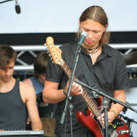 Two male musicians performing on stage. The foreground musician, with long brown hair, a black shirt, and playing a red electric guitar while singing into a microphone, exudes the rock vibes of 2011. The background musician has short dark hair, a black tank top, and is playing a Laba keyboard. Drums are partially visible.
