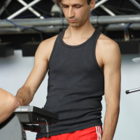 A person wearing a black tank top and red athletic shorts with white stripes is standing and focused on using an electronic device on stage. They have short, dark hair and are surrounded by stage equipment, including metal trusses and microphones in the background, reminiscent of a 2011 concert.