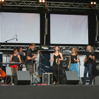 A band performing on an outdoor stage features nine musicians playing instruments including cello, violin, electric guitar, and bass. Some musicians are seated while others stand. They are surrounded by equipment such as microphones, stands, and speakers, with blank projection screens in the background. Laba daba 2011 vibes!