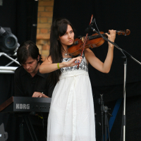 A woman in a white dress plays a violin on stage, concentrating on her music. Behind her, a man dressed in black plays a Korg keyboard. The dark background reveals stage equipment and a brick wall. Both musicians appear focused on their performance at the 2011 Laba Daba festival.