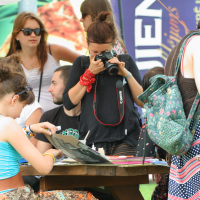 A group of people are gathered outdoors at Laba Daba 2011. One person is using a Canon camera to take a photo, while others sit at a table engaged in crafts and drawing. The atmosphere is casual and collaborative, with various outfits and accessories, including hats, sunglasses, and backpacks.