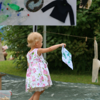 A young child with blonde hair, wearing a white dress with pink flowers, stands on grass and holds up a colorful drawing. Behind her, a sign with Latvian text and a collage are visible. The scene appears to be an outdoor daba event or activity in a garden or park with trees and greenery in the background.