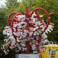 A red metal sculpture in the shape of a tree, created for the 2011 Laba Daba festival, holds numerous attached plastic bottles. Positioned outdoors with lush green trees in the background, it stands next to a yellow recycling bin and a silver ladder, with a sign at its base encouraging plastic bottle recycling in Latvian.