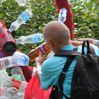 A person with cropped hair, wearing a turquoise shirt and carrying a black backpack, places a plastic bottle into a recycling sculpture made of numerous plastic bottles and other items at the Laba Daba 2011 festival. The background features green foliage and blurred outdoor scenery.