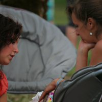 Two women are seated outdoors on foldable chairs. One woman, wearing an orange shirt and white gloves, is carefully applying a bandage to the other woman's hand, who is wearing a colorful floral dress from 2011. Both are deeply focused on the task. The background shows a grassy area and blurred people.