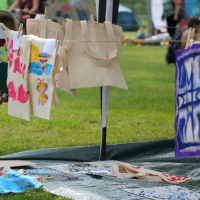 Fabric tote bags with colorful hand-painted designs, including abstract patterns and a butterfly, hang to dry on a clothesline at the 2011 Laba Daba outdoor arts and crafts fair. A green grassy field and people seated in the background are slightly blurred. Some art supplies are scattered on the ground below.