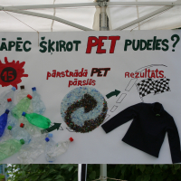 A poster displayed under a white tent at Laba daba 2011 illustrates the benefits of recycling PET. It has text in Latvian and a visual flow showing crumpled plastic bottles transforming into plastic pellets and then into a black fleece jacket, with a racing flag next to it representing the end result.