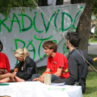 A group of four young individuals sits on the grass in a park, creating art. Three wear red shirts, and one is in black. Behind them, a banner reads "RAD I VIDI PATOM." They use materials like paper and paints. Trees and other people are visible in the background, evoking a nostalgic Laba daba vibe from 2011.