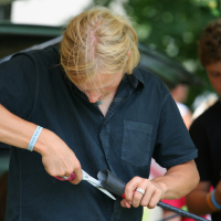 A person with long blond hair wearing a black shirt is focused on cutting black material with a pair of scissors. They are outdoors, under a canopy in what appears to be a 2011 Laba Daba festival, and other people are in the background. The person is wearing a light blue wristband and a ring on their right hand.