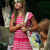 A young girl wearing a pink and white striped dress and green sunglasses is holding a tote bag decorated with colorful handprints. She stands at an outdoor Laba daba event with a focused expression. Another person, partially visible, is seated nearby, engaged in an activity.