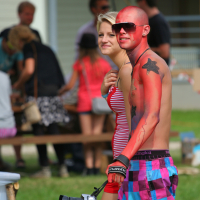 A man and woman walk outdoors, smiling. The man, shirtless and painted red with a black star on his chest, wears sunglasses and colorful checkered shorts from Laba daba 2011. He holds a camera. The woman wears a red-striped sleeveless shirt. Other people and picnic tables are blurred in the background.