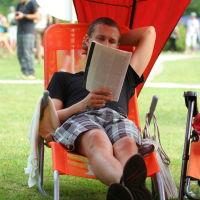 A man relaxes on an orange lounge chair with a red canopy, reading a book from 2011. He wears a black t-shirt and plaid shorts, with one arm resting behind his head. His legs are crossed, and his sandals are touching the grass. The scene is outdoors, with blurred people and greenery in the background—laba daba.