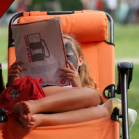 A child with face paint lies on an orange chair outdoors, reading a 2011 magazine. The cover features a gasoline pump and text in a non-English language. The child's legs are crossed and relaxed, while the background shows grassy area and blurred figures, creating a tranquil Laba daba scene.