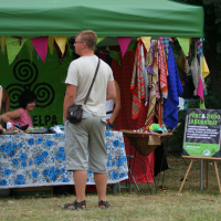 A man in a white shirt and beige shorts stands in front of a green tent with colorful triangular flags, reminiscent of Laba Daba 2011. Two women behind a table with a blue floral cloth are selling goods. To the right, a rack of clothes is displayed. A nearby sign contains text, and a leafy tree is in the background.