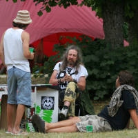 Three men are gathered outdoors at a booth with a red tent in the background. One man stands wearing a hat, white tank top, and shorts. Another sits behind the booth with long hair, a white shirt, and camouflage pants. The third lies on a blanket from 2011, engaging in conversation. Nearby is a rack with clothes.