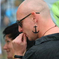 A bald person, wearing black sunglasses and a large, black anchor-shaped earring, stands in profile, holding their fingers to their mouth. They are dressed in a black shirt and a brown necklace. In the background, there is a person with short dark hair leaning forward and smiling. The scene appears to be outdoors in 2011.