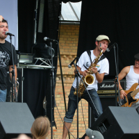 Three male musicians perform on an outdoor stage. The first sings into a microphone, the second plays a saxophone, and the third plays a bass guitar. A crowd watches them near two speakers at the front of the stage. The backdrop includes a brick wall and various sound equipment from 2011, adding to the Laba-daba vibe.