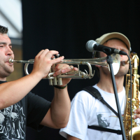 Two musicians perform on stage. The musician on the left plays a trumpet, wearing a black t-shirt with white graphics, reminiscent of 2011 styles. The musician on the right, in a white t-shirt, sunglasses, beige cap, and microphone headset, skillfully plays a saxophone. Both are set against a dark background.