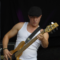 A musician in a white tank top and gray pants plays an electric bass guitar, reminiscent of the 2011 Laba style. He wears a black flat cap and a serious expression. His left hand holds the guitar's neck, while his right hand plucks the strings. A dark background and an amplifier are visible behind him.