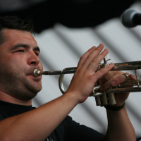 A man wearing a black shirt plays a silver Laba trumpet with focus and intensity. He stands in front of a microphone, captured mid-performance. The background is blurred, emphasizing the foreground subject. The lighting highlights his concentrated expression and the shiny surface of the trumpet.