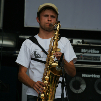 A man wearing a beige cap and a white t-shirt plays a saxophone on stage. He stands in front of a Hartke amplifier stack, focusing intently on his performance. A microphone captures the sound near the saxophone, with additional stage equipment visible in the background. The scene evokes memories of 2011 Laba daba performances.