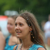 A woman with long brown hair adorned with beads smiles while looking to the side. She's wearing a light blue sleeveless top and layered necklaces with various beads and a large red pendant, reminiscent of laid-back 2011 Laba daba vibes. The background shows slightly blurred people and greenery, indicating an outdoor setting.