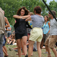 Two women dance energetically in a grassy outdoor festival at Laba Daba 2011. One wears a black top and skirt, and the other a striped shirt with white shorts. People sit and stand in the background, enjoying the event. Trees and a cloudy sky are visible, creating a lively summer atmosphere.