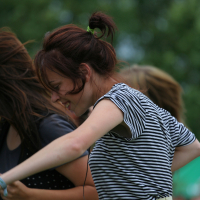A young woman with dark hair styled in a messy bun is dancing energetically outdoors, reminiscent of a lively scene from 2011. She is wearing a black and white striped shirt and light-colored pants. The background shows blurred foliage, suggesting a park or garden setting. Others are partially visible around her, also engaged in the activity.
