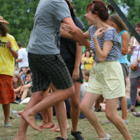 A group of young people are dancing and having fun outdoors on a grassy area. A young man in a gray T-shirt and plaid shorts is enthusiastically dancing with a young woman in a striped shirt and light-colored shorts. It's 2011, and other people in casual summer clothes are watching and enjoying the scene under the trees.