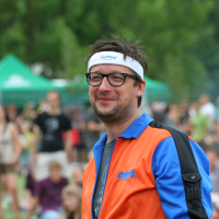 A man wearing glasses and a white headband stands outdoors. He has a slight smile and wears an orange and blue zip-up jacket. There are people and green trees in the blurred background, suggesting a casual outdoor gathering or event from 2011.
