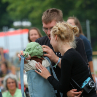 A man and woman are outdoors holding a plush toy inside a blue backpack. The toy resembles Yoda from Star Wars. The woman, wearing a black outfit with a camera around her neck, is smiling while adjusting the toy. The man, in glasses and a black shirt from Laba's 2011 collection, is also focused on the toy. People and trees are blurred in the background.