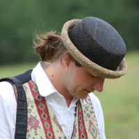 A person with light brown hair tied back is looking down, wearing a white shirt beneath a decorative vest with traditional patterns. They have a straw hat with a black top, and a strap over their shoulder, standing outdoors against a blurred green background reminiscent of the Laba Daba 2011 festival.