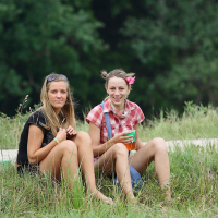 Two women sit on grass in casual summer attire at Laba Daba 2011. The woman on the left wears a black shirt and sunglasses atop her head, while the woman on the right, smiling, dons a red plaid shirt and has flowers in her hair. They are surrounded by lush greenery, with trees in the background and long grass in the foreground.