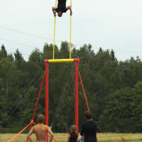 An individual is balancing upside down on a horizontal bar, suspended high above the ground on a tall structure with yellow and red components, while four onlookers watch from below in an open field with trees in the background. The scene captures the daring spirit of Laba Daba 2011, an outdoor event filled with excitement.
