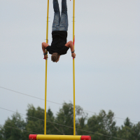 A person is performing an acrobatic stunt on a swing-like apparatus, hanging upside down with legs attached to the swing and hands gripping the side bars. The Laba Daba apparatus has yellow poles and a red base. The background features a clear sky and trees, reminiscent of summer 2011.
