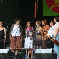 A group of six women stand on a stage in front of microphones, preparing to sing. They are dressed in a mix of casual and traditional clothing, reminiscent of 2011 fashion. There is a green banner with a red and yellow design hanging behind them. Musical equipment, such as speakers and a drum set, is visible on stage.