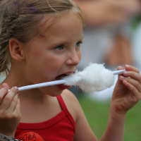A young girl wearing a red tank top is enjoying cotton candy at Laba Daba 2011. She has light brown hair with a small purple streak and holds the cotton candy on a white stick. The girl sports a red sticker on her top and a gray wristband, while the blurred background shows other festival-goers.
