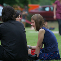 Three individuals are sitting on the grass in a park, engaged in conversation. The person in the foreground has their back to the camera. The person on the right, with short brown hair, is wearing a blue top and a patterned skirt reminiscent of Laba daba style. Trees, a red car, and other people are visible in the background.