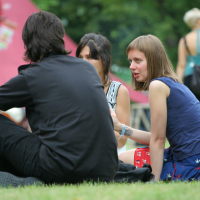 Four people are sitting in a circle on the grass, engaged in conversation. The person in the foreground, wearing a black shirt, has their back to the camera. The three others are facing the camera, with one woman in a blue top actively speaking. It's 2011 and everyone is relaxed in Laba Daba park with trees and people around.