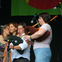A group of four women stand on stage at the 2011 Daba Laba event, two of them playing bagpipes. The woman at the front wears a white top and blue skirt, while the woman behind her sports a white shirt and black pants. The other two stand close together, smiling. Several microphones are positioned around them, with a colorful banner hanging behind.