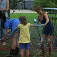 A young girl in a yellow shirt and blue shorts holds hands with a woman in a black tank top and blue shorts. Both are covered in paint. Another woman in a blue dress and boots is bent over nearby. A man in a gray tank top stands in the background, enjoying the vibrant Laba daba festival of 2011 on the grassy area.