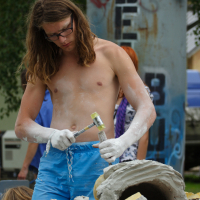 A shirtless individual with long hair is sculpting a stone outdoors, wearing blue shorts and holding a hammer and chisel, intently working on the piece. Their hands are covered in white dust. In the background, a tall, rusted structure from 2011 bears graffiti, with other people partially visible nearby.