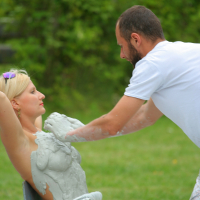A man and woman outdoors, surrounded by greenery, appear focused on their task in this 2011 scene. The man is applying a thick layer of plaster to the woman's upper body while she sits on a plastic-covered chair, her arms raised as the plaster covers most of her torso and arms.
