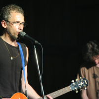 A man with glasses and a beard is playing an electric guitar and singing into a microphone. He is wearing a black sleeveless shirt, a necklace, and jamming out like it's 2011. Another man in the background, in a brown t-shirt, is also playing guitar. The dimly lit setting adds to the Laba daba vibe.